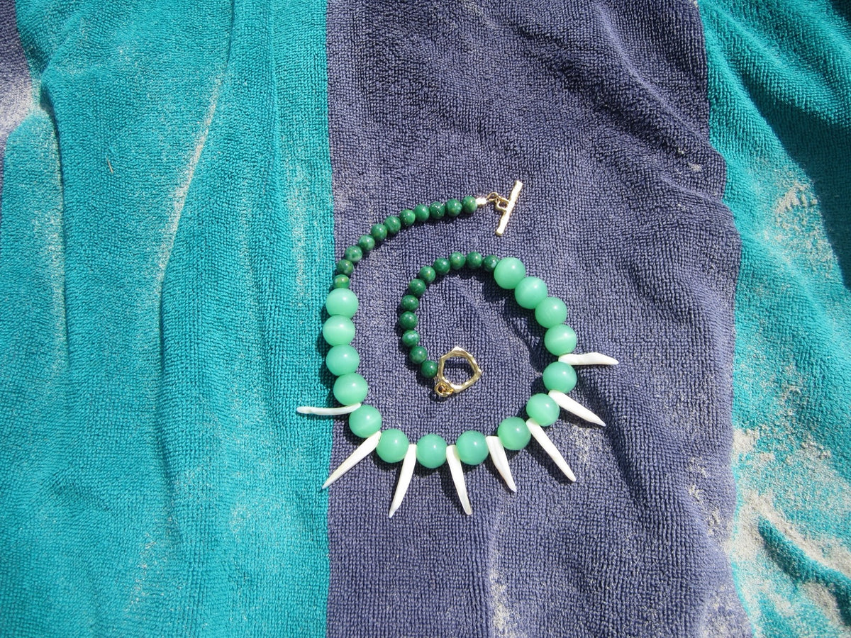 A green beaded necklace with white shark teeth accents and gold-tone hardware, laid out on a blue and teal towel.