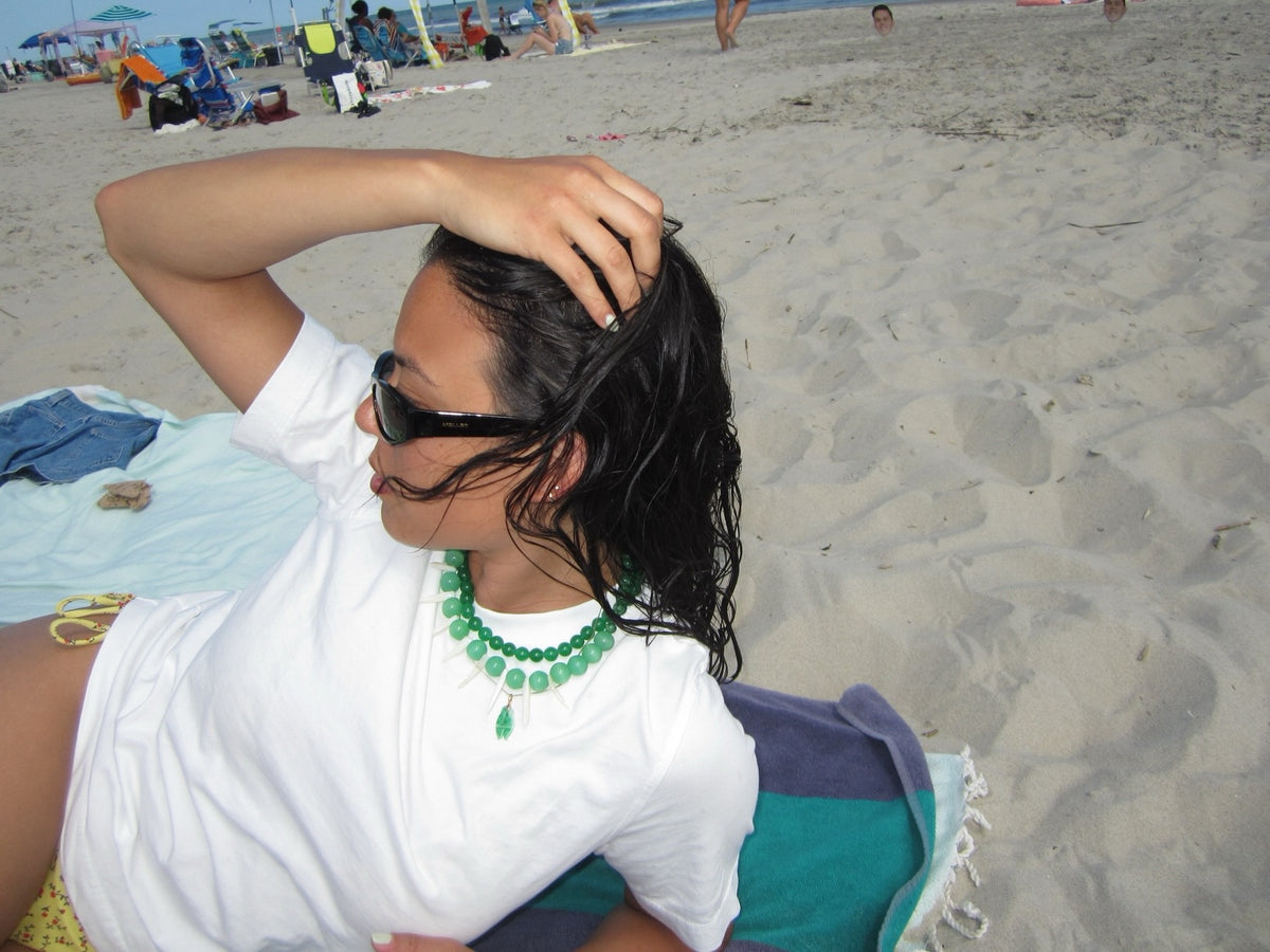 A woman with wet dark hair sits on a beach towel on a sandy beach, wearing a white t-shirt, black sunglasses, and a chunky green beaded necklace.