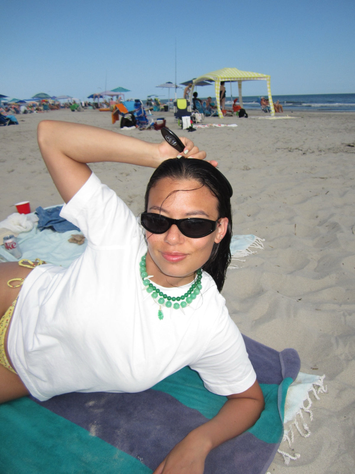 A woman wearing a white t-shirt and a green beaded necklace lies on a beach towel on a sandy beach, holding a hairbrush to her hair under a clear blue sky.